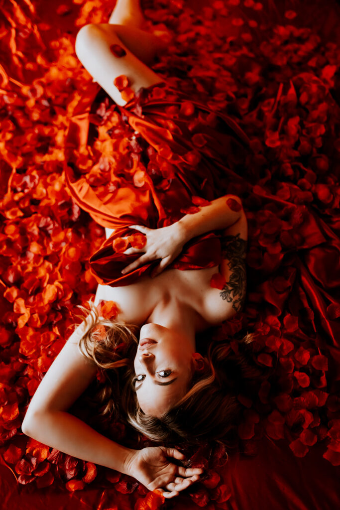 "Luxurious boudoir setting on a red silk bed covered in thousands of red rose petals."
"Sensual moment captured on a velvety red silk bed adorned with a cascade of red rose petals."
"Elegant and romantic boudoir scene featuring a bed covered in red silk and a blanket of rose petals."